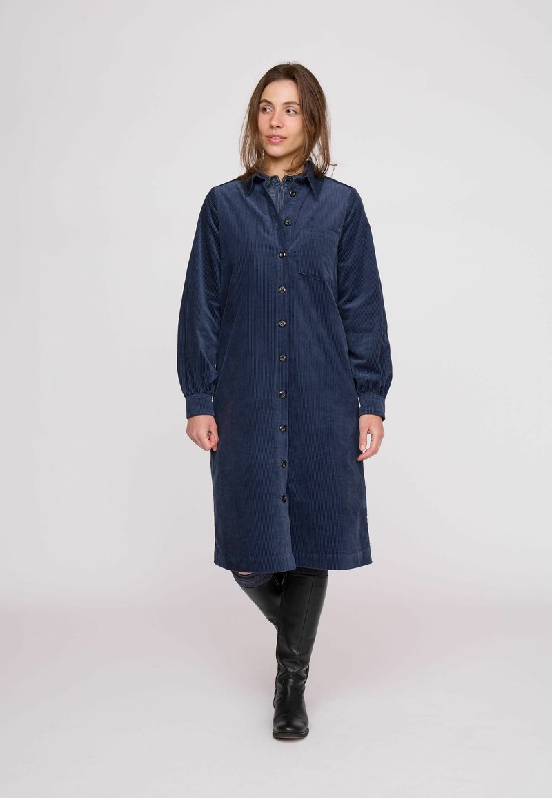 Námornícky modré šaty z kordurovej látky s predným zapínaním na gombíky, dlhými rukávmi, golierom a náprsným vreckom. Tkanina má rebrovanú textúru a siaha pod kolená.