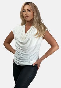Biała, drapowana bluzka z dekoltem w kształcie stójki i krótkimi rękawami. Teksturowany materiał, dopasowany krój. Noszona z czarnymi spodniami z dekoracyjnymi guzikami.