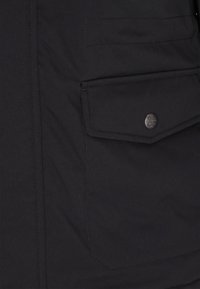 Fekete dzseki sima, matt felülettel, nagy fedeles zsebbel, ezüst gomb hardware-rel és látható varrási részletekkel.