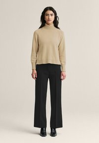 Khaki sweateri, jossa on korkeakaulus ja ribbineule, yhdistetty mustiin leveälahkeisiin housuihin ja mustiin nilkkureihin, seisoo neutraalia taustaa vasten.