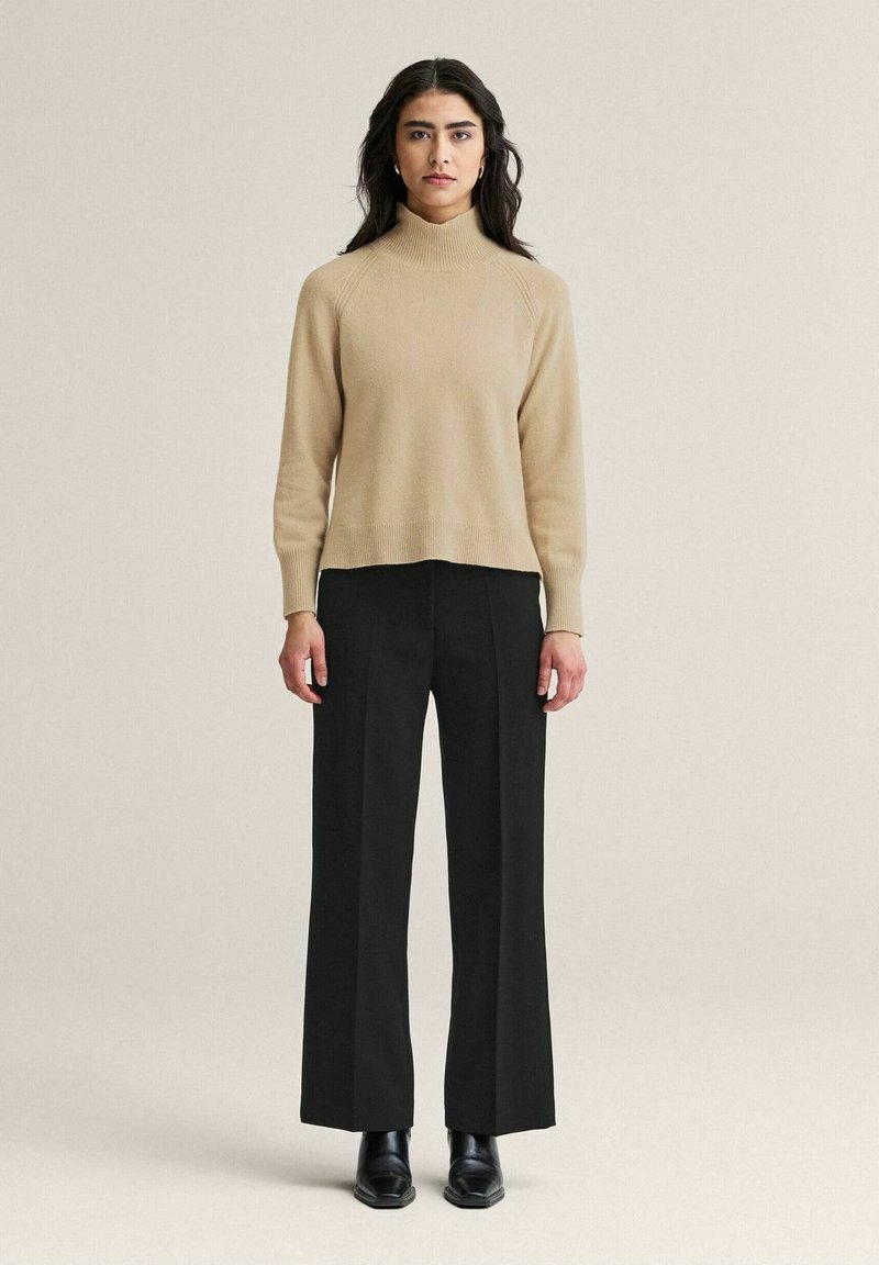 Khaki sweateri, jossa on korkeakaulus ja ribbineule, yhdistetty mustiin leveälahkeisiin housuihin ja mustiin nilkkureihin, seisoo neutraalia taustaa vasten.