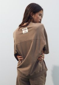 Smeđi oversized dukserica s oznakom na stražnjoj strani. Mekana tekstura, kratki rukavi i opušteni kroj. Nošena uz usklađene hlače i minimalne dodatke.