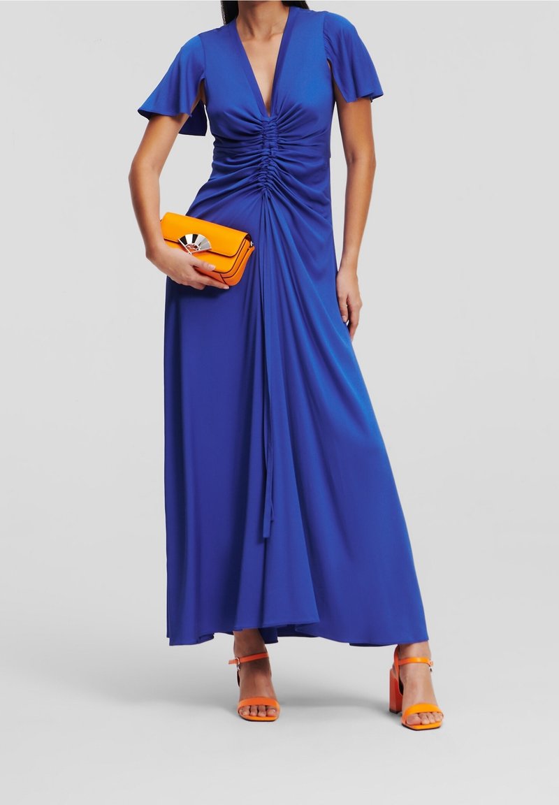 Kvinna i en lång blå dräkt med rynkor, som håller en orange clutchny och bär orange högklackade sandaler, stående mot en enfärgad bakgrund.