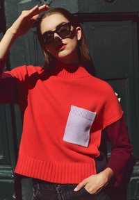 Rode gebreide trui met een hoge ribbelkraag, voorzien van een paarse zak en contrasterende donkere mouwen; met een textuurstof en ribbelzoom.