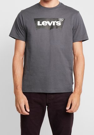 Homme portant un t-shirt gris Levi's avec logo noir et pantalon foncé, debout les bras détendus le long du corps.