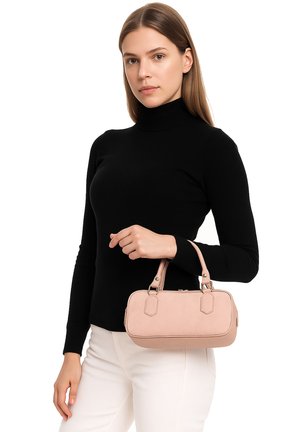 Giovane donna con dolcevita nero e pantaloni bianchi che tiene una piccola borsa beige con doppi manici su sfondo bianco.