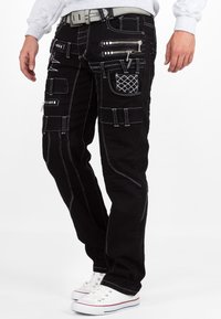 Pantaloni cargo neri in denim con cuciture bianche. Presentano numerose tasche, cerniere e un motivo testurizzato. Indossati con sneakers bianche.