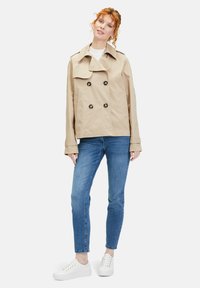 Beige double-breasted trench coat with oversized collar, black buttons, and side pockets; paired with blue jeans and white sneakers.