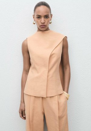 Pfirsichfarbener ärmelloser Top mit asymmetrischem Design, kombiniert mit passenden Hosen. Der Stoff wirkt glatt mit einer feinen Textur. Goldene Creolen.