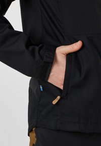 Hand insatt i sidofickan på en svart jacka med en liten svensk flaggetikett och kardborrbandsdetalj på ärmen.