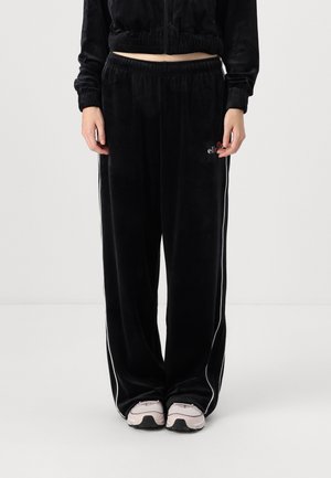 Personne portant un pantalon de survêtement large en velours noir avec des bandes blanches sur les côtés et des baskets rose clair, debout sur un fond blanc uni.