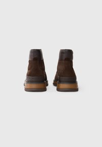 Botas de tobillo de ante marrón con un suave collar de cuero marrón oscuro y una gruesa suela de goma marrón claro. Presenta un sutil logotipo en el lateral.