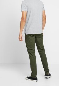 Muž v olivově zelených kalhotách, světle šedém tričku s krátkým rukávem a černých nízkých teniskách stojí proti jednoduchému bílému pozadí.