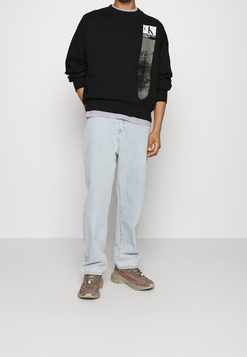 Homme portant un sweat-shirt noir Calvin Klein, un jean bleu clair et des baskets marron, debout devant un fond clair uni.