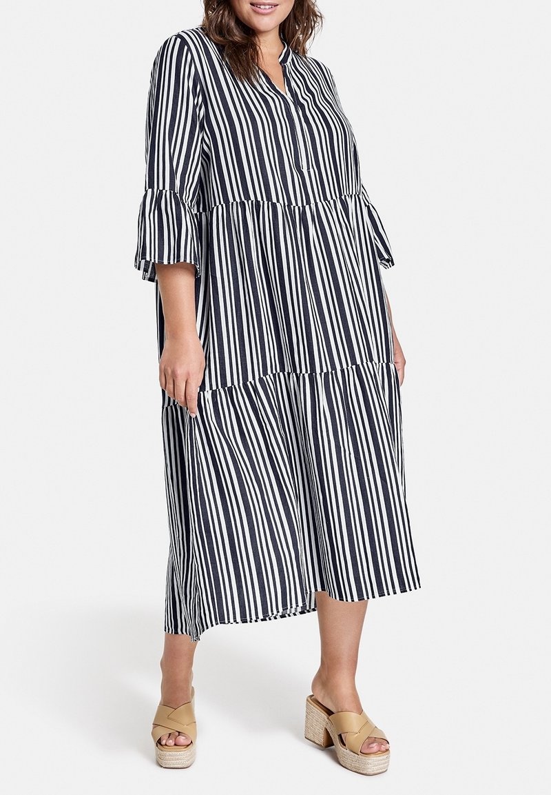 Femme portant une robe midi à rayures verticales noires et blanches avec des manches 3/4 à volants et des sandales compensées beiges, debout sur un fond blanc uni.