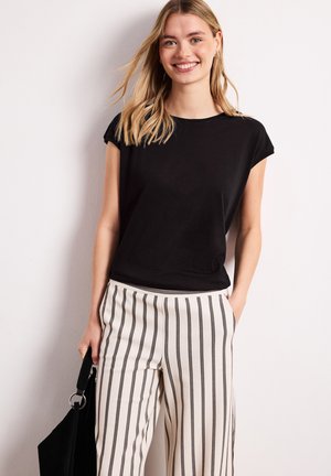 Jeune femme sourit, portant un haut noir sans manches et un pantalon blanc avec des rayures verticales noires, tenant un sac noir avec une main dans la poche.