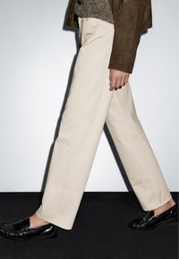 Béžové kalhoty na míru se rovnými nohavicemi, vyrobené z bavlněné tkaniny, v kombinaci s černými lesklými polobotkami, vyzdvižené na neutrálním pozadí.