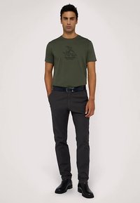 Olivově zelené bavlněné tričko s černým grafickým motivem na přední straně, doplněné o tmavě šedé úzké kalhoty a černé kotníkové boty.