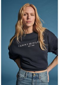 Русокоса жена, облечена с тъмносиня блуза Tommy Hilfiger и сини дънки, стояща с ръце на ханша на син фон.