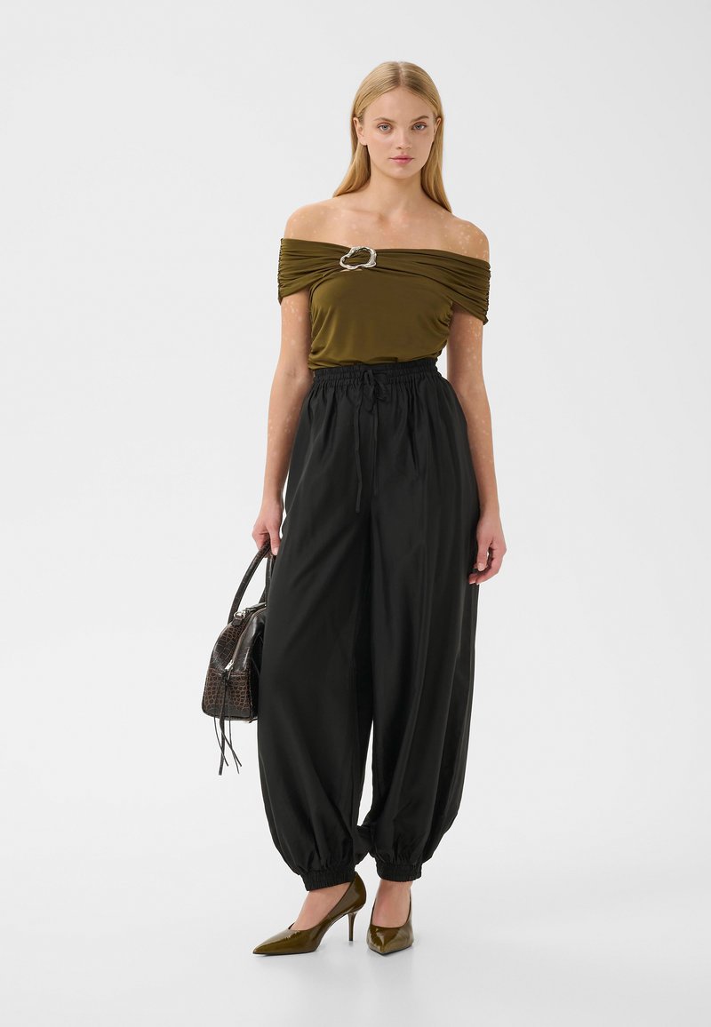 Femme portant un haut vert olive épaules dénudées avec une boucle argentée, un pantalon ample noir taille haute, des talons olive, tenant un sac à main marron foncé.