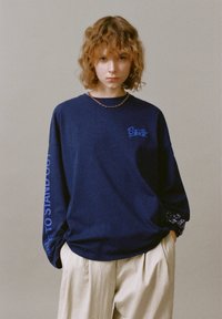 Felpa a maniche lunghe blu navy con testo grafico, caratterizzata da una vestibilità rilassata e polsini arrotolati che mostrano un motivo a quadri. Pantaloni beige a vestibilità rilassata.