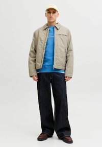 Bežinė užtrauktuku striukė su šonine kišene, uždėta ant mėlynos sportinės palaidinės. Palaidi tamsūs džinsai ir rudi batai užbaigia aprangą.