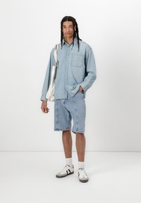 Cămașă din denim de culoare albastru deschis, cu mâneci lungi și un buzunar pe piept, asortată cu pantaloni șort din denim. Tenisi negri și albi completează look-ul.