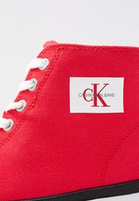 Červené plátěné vysoké tenisky s bílými tkaničkami, kovovými očky a bílým štítkem s nápisem "CALVIN KLEIN JEANS" a červeným logem "CK".