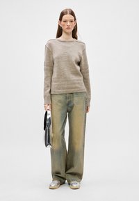 Beige sweater with textured sleeves, paired with loose faded jeans. Model holds a shiny silver bag, wearing metallic shoes. Plain background.