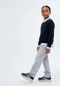 Pulover bleumarin peste o cămașă cu dungi albastre deschise, blugi gri deschis și teniși multicolori cu accente bleumarin, verde și bej.