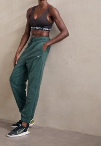 Idrottslig person i svart sportbhn, gröna Nike träningsbyxor och svarta sneakers står mot en beige vägg med en hand i fickan.