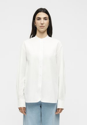 Γυναίκα με ίσια σκούρα μαλλιά φοράει ένα λευκό πουκάμισο με μακριά μανίκια και ανοιχτά μπλε τζιν, στέκεται μπροστά σε ένα απλό λευκό φόντο.