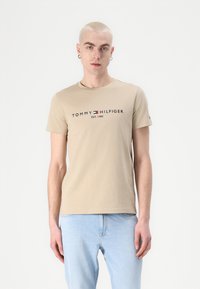 Bežinė medvilninė marškinėliai su apvalia iškirpte, su juodu logotipo tekstu "TOMMY HILFIGER EST. 1985" ir mažesniu logotipo akcentu ant rankovės.
