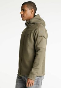 Maslinasto zelena zip-up dukserica s teksturiranim završetkom, podesivim kapuljačom, bočnim džepovima i logotipom na grudima, prikazana na modelu.
