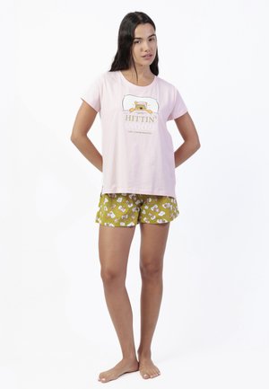 Mujer vestida con una camiseta de manga corta color rosa claro con el texto "Hittin' Snooze" y pantalones cortos verdes para dormir con un patrón de personajes de dibujos animados, de pie y descalza.
