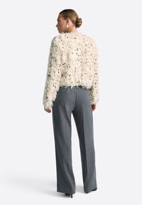 Textured cream jacket with fringe details, paired with high-waisted gray trousers. The outfit features a relaxed fit and subtle sheen.