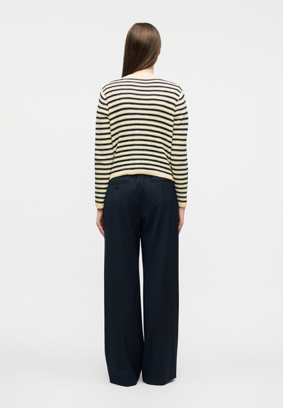 Γυναίκα με μακριά ίσια μαλλιά, φορώντας ένα πουλόβερ με ρίγες σε κρεμ και ναυτικό χρώμα και παντελόνι μαύρο, φαρδύ, που στέκεται γυρισμένη με την πλάτη σε απλό φόντο.