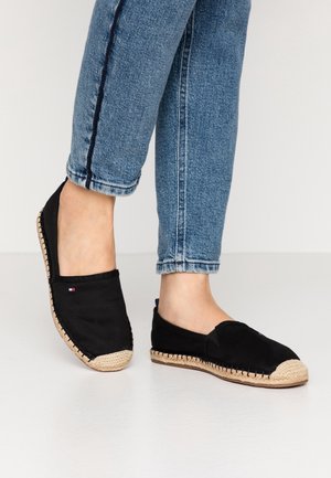 Henkilö, jolla on mustat espadrillot kudotulla beigen värisellä pohjalla ja ylös käännetyt siniset farkut, seisoo vaalealla lattialla.