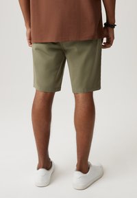 Oliwkowe szorty o gładkiej fakturze, prostym kroju i podwiniętych nogawkach, w zestawieniu z białymi sneakerami na nagich nogach.