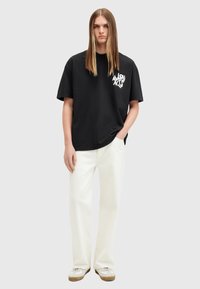 Camiseta de algodón negra con un diseño gráfico blanco, combinada con pantalones blancos y zapatos beige, sobre un fondo neutro.