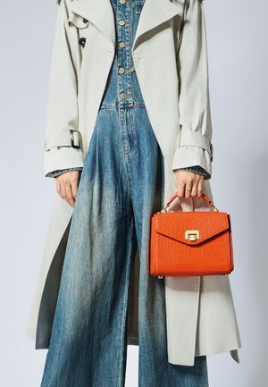 Borsa arancione in pelle effetto coccodrillo con un design strutturato, accessori in oro e manico superiore, abbinata a un trench coat beige e un outfit in denim.
