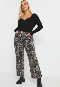 Nainen seisoo kädet leopardikuvioisten housujen taskuissa, päällään musta istuva pitkähihaine paita ja mustat nilkkurit valkoista taustaa vasten.