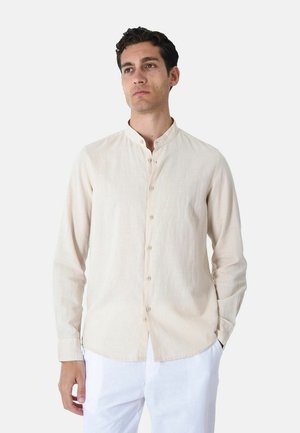 Uomo che indossa una camicia beige a maniche lunghe con colletto alla mandarina e pantaloni bianchi, in piedi con una mano in tasca, guardando leggermente di lato.