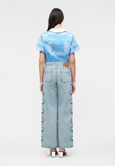 Γυναίκα με μακριά σκούρα μαλλιά που φορά μπλε πουκάμισο με σχέδιο μπαντάνας, ανοιχτόχρωμο τζιν παντελόνι φαρδύ με πλαϊνές λεπτομέρειες και λευκά σανδάλια με χοντρό τακούνι.