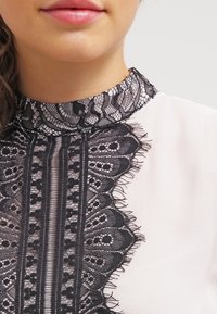 Bílá blůza s vysokým černým krajkovým límcem s jemnými vzory a třásněmi, které vytvářejí texturovaný kontrast u výstřihu.