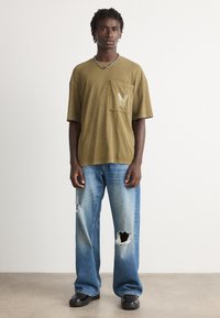 Person står iført en olivenfarvet oversized T-shirt med lommer foran, slidte vide blå jeans, sorte sko og en sølvfarvet kædehalssmykke foran en ensfarvet baggrund.