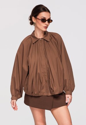 Kvinde med mørkt hår sat op i en knold iført brun oversized plisseret jakke, matchende mininederdel, solbriller og guldringe mod ensfarvet baggrund.