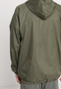 Chaqueta con capucha de color verde oliva, con un acabado ligero y suave. Presenta un dobladillo elástico y un corte relajado, diseñada para la comodidad y facilidad de movimiento.