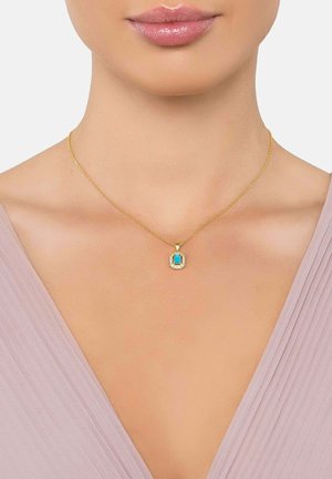 Collar de oro con un colgante rectangular de turquesa rodeado de pequeños diamantes, llevado en el cuello de una mujer con una blusa de cuello en V de color púrpura claro.