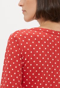 Czerwony prążkowany top w białe kropki, posiada okrągły dekolt i krótkie rękawy. Materiał jest miękki, a sylwetka dopasowana.